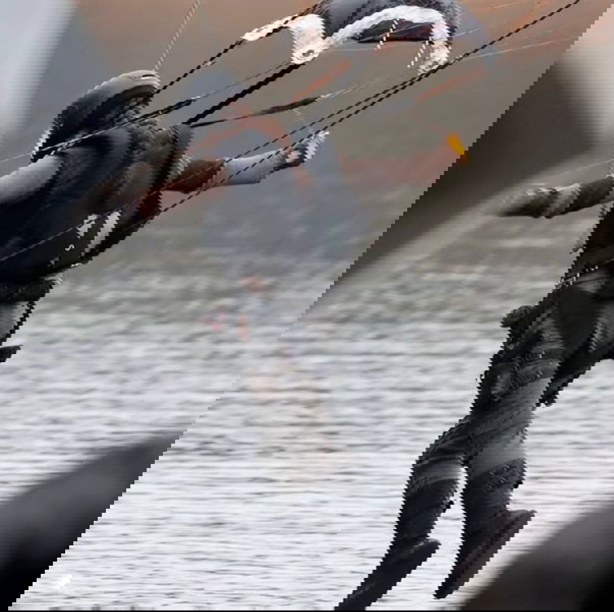 Камера фиксирует сцену: актриса с косой и защитным шлемом спускается к воде с парашютом, видно снаряжение и тросы.