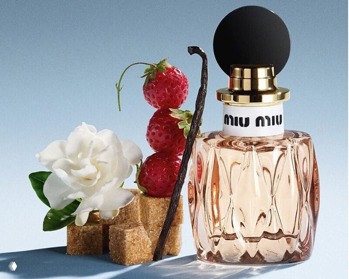 Флакон парфюма Miu Miu на светлом фоне рядом с ягодами земляники, гарденией, палочкой ванили и кубиками сахара.