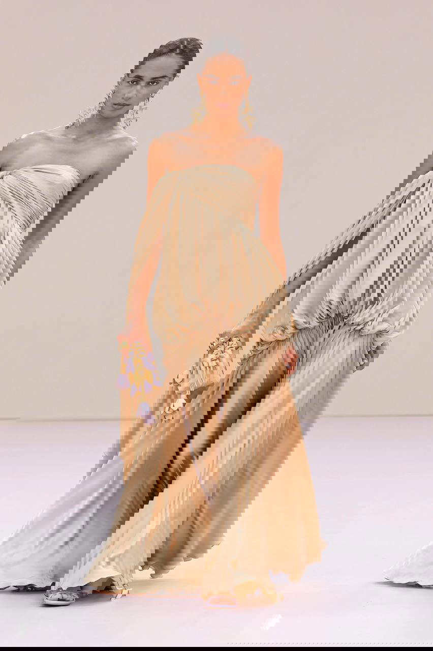 Модель на подиуме в бежевом платье с античными мотивами: асимметрия на одно плечо, мягкие складки и летящий шелк, образ для торжеств и фотосессий.