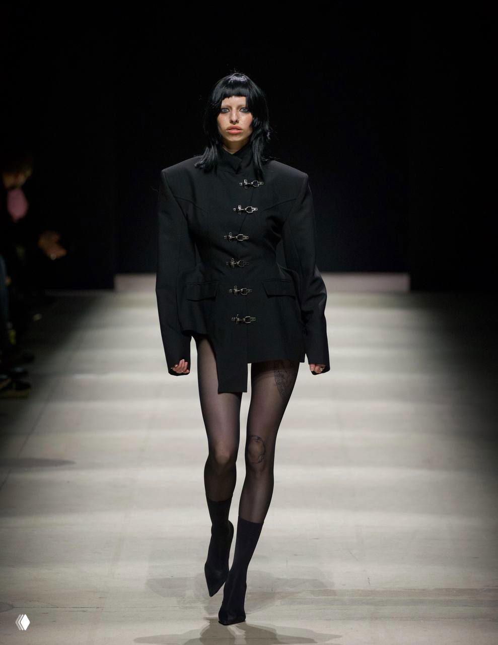 Показ с акцентом на мужские плечи: модель в черном жакете с широкой плечевой линией и прозрачными колготками на подиуме.