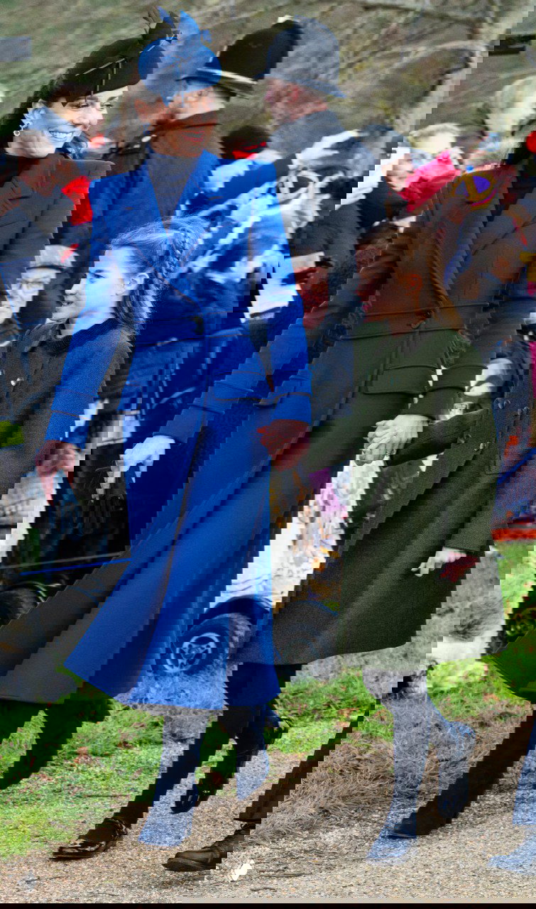 Женщина в ярком синем пальто и шляпке держит за руку ребёнка в зелёном пальто на прогулке во время рождественского события, вокруг публики.