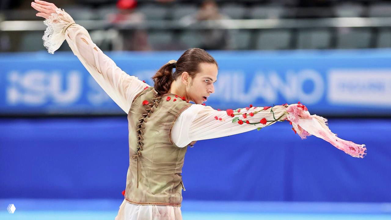 Панорамный кадр проката: фигурист в белой блузе и коричневом жилете выполняет па в эффектном наряде с розами.