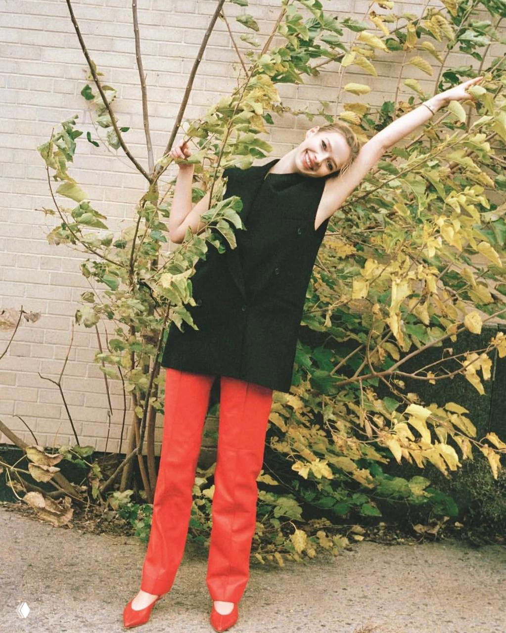 Уличный кадр молодой модели в чёрном жилете и ярко-красных брюках среди жёлтой листвы — лёгкая и непринуждённая поза в fashion-стилистике.