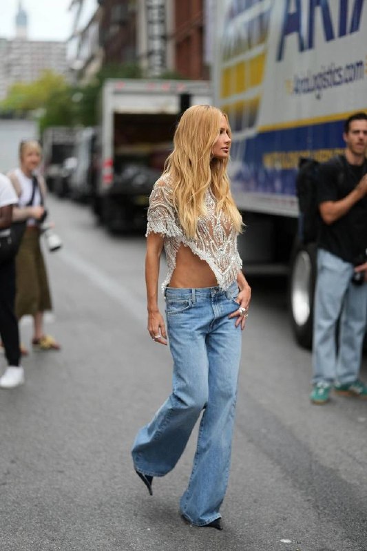 Уличный кадр: блондинка в украшенном прозрачном топе и широких джинсах с низкой посадкой прогуливается по городу в стиле стритстайл.