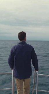 Мужчина в синей куртке стоит спиной на корме лодки и смотрит на море и облачное небо — атмосферный кадр из кампании.