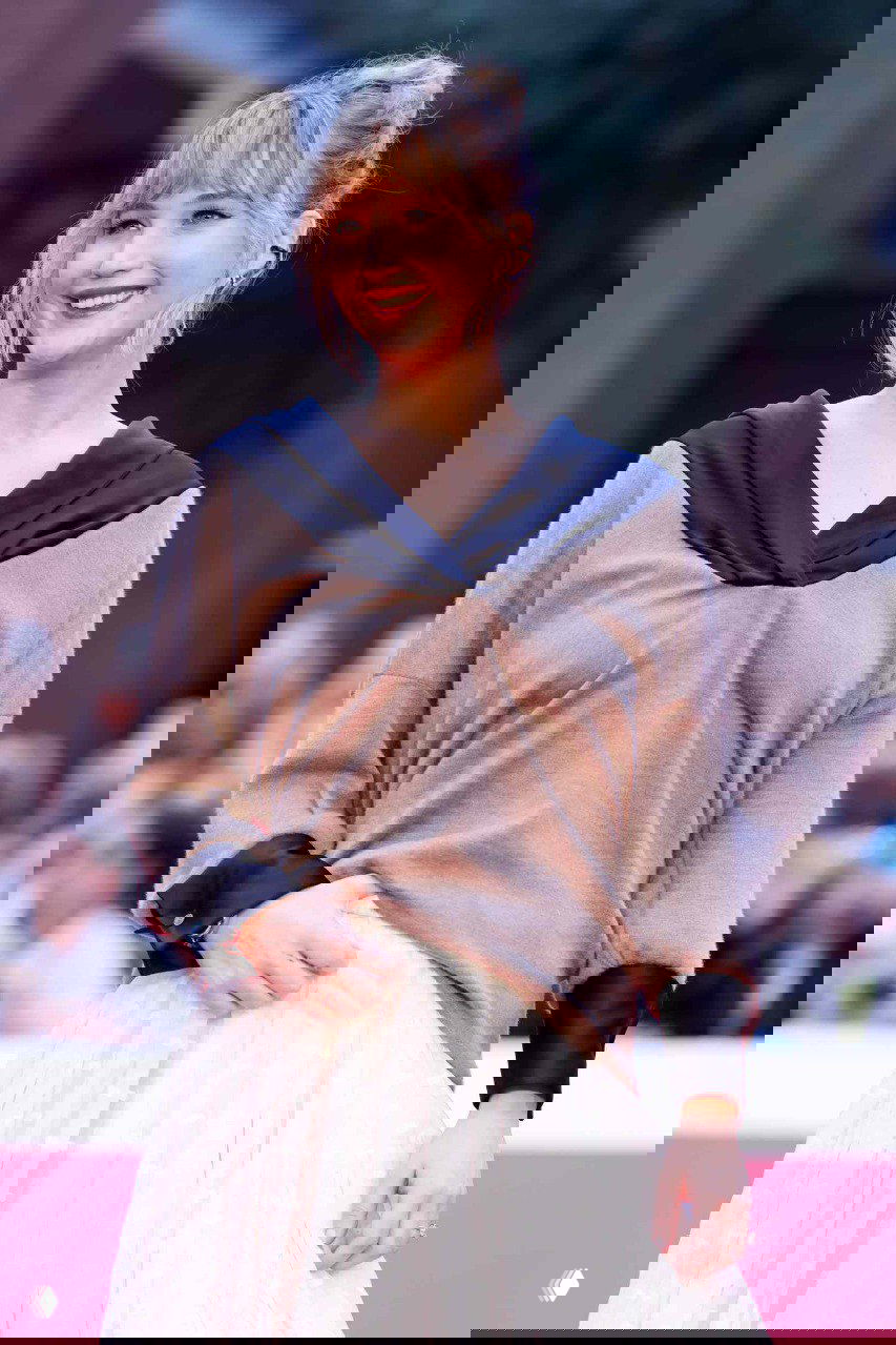 Портретный снимок с улыбкой, где видно верх образа — мягкий коричневый свитер с чёрным V‑образным воротником и пышная светлая юбка.