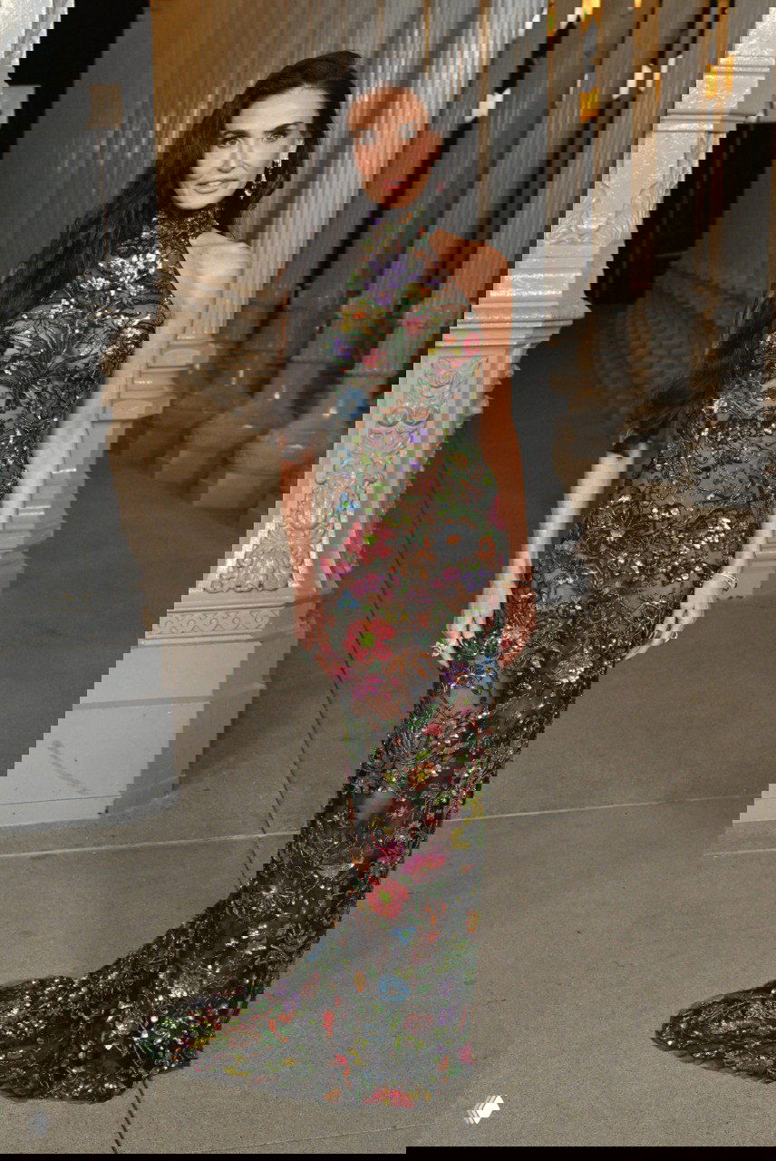 Женщина в полупрозрачном платье с цветочной вышивкой и длинным шлейфом позирует у колонн инсталляции, яркий декоративный узор.