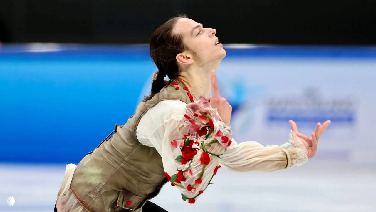 Фигурист с поднятой головой и выразительной постановкой рук в костюме с лепестками роз на фоне ледовой арены.