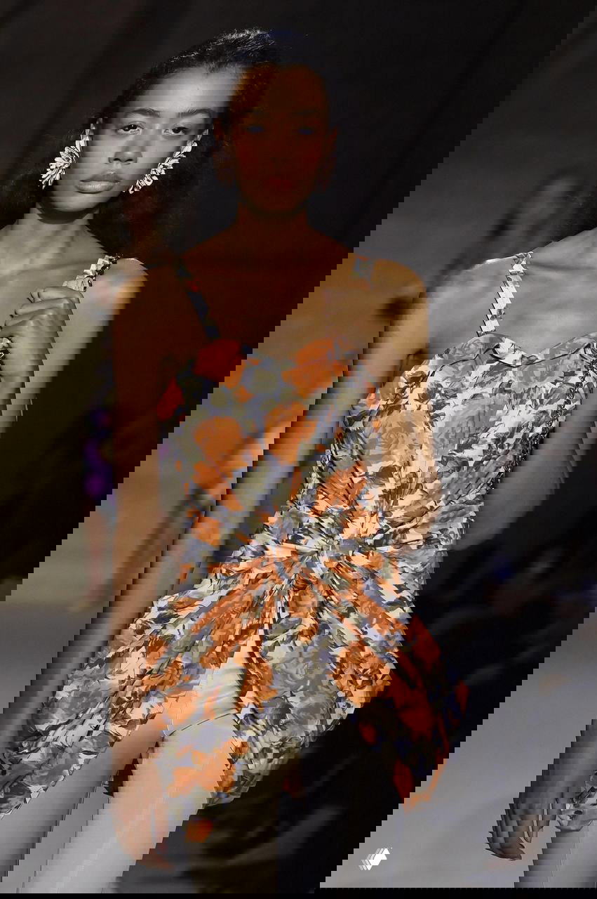 Модель в лёгком платье с крупным оранжевым цветочным узором, акцент на текстуру и складки ткани в кадре с модного показа.