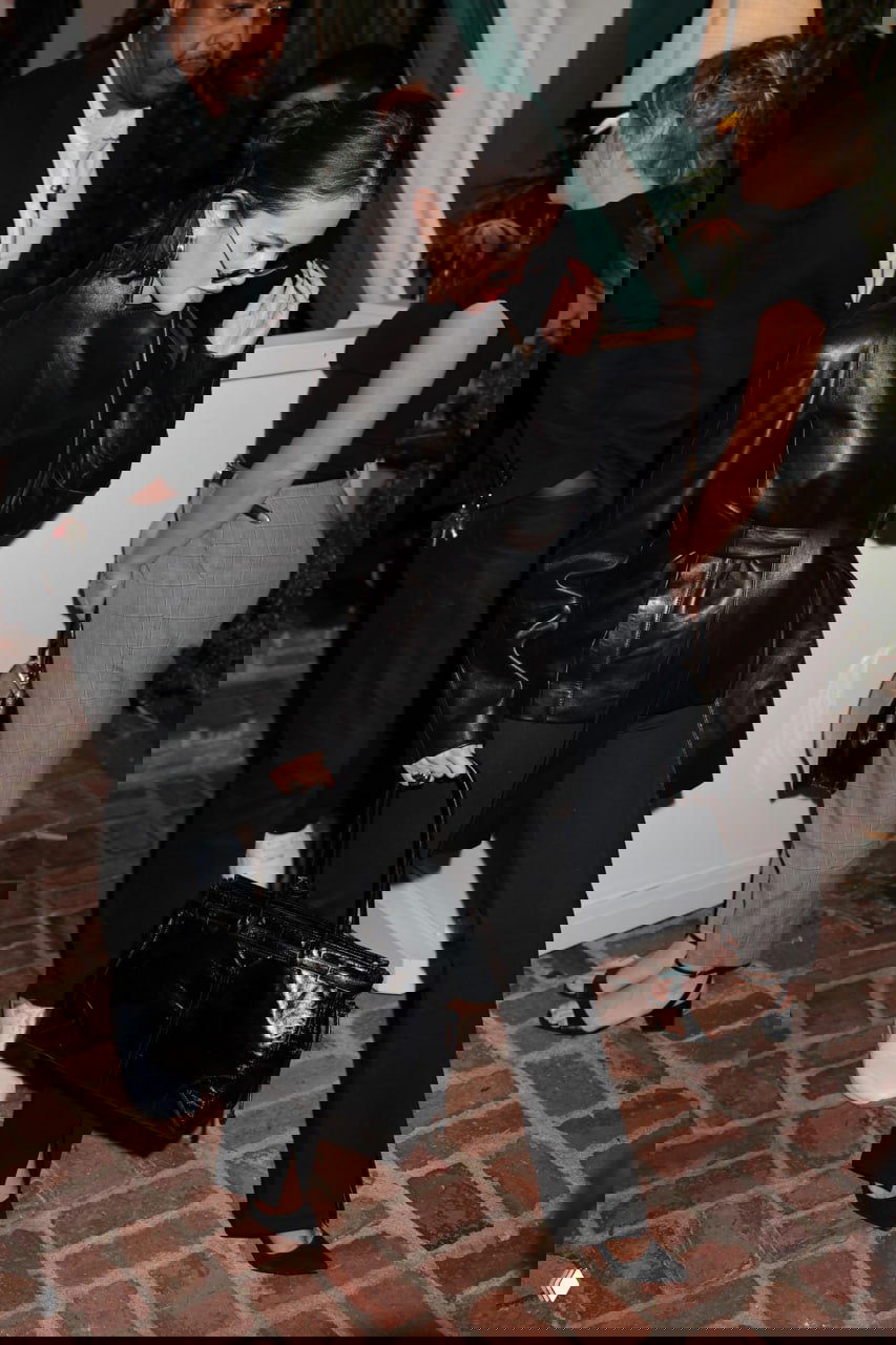 Женщина в чёрной кожаной куртке и прямых брюках идёт по вечерней улице с большой чёрной сумкой рядом с другими людьми. Фотография уличного папарацци.