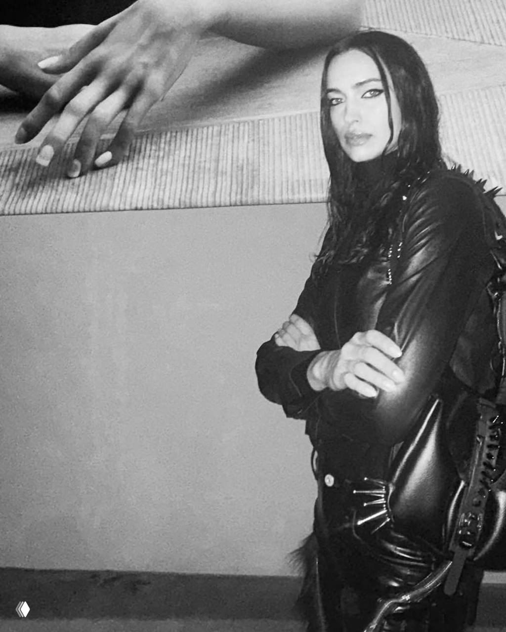 Чёрно‑белый кадр: модель в кожаной куртке со скрещёнными руками на фоне крупного художественного принта.