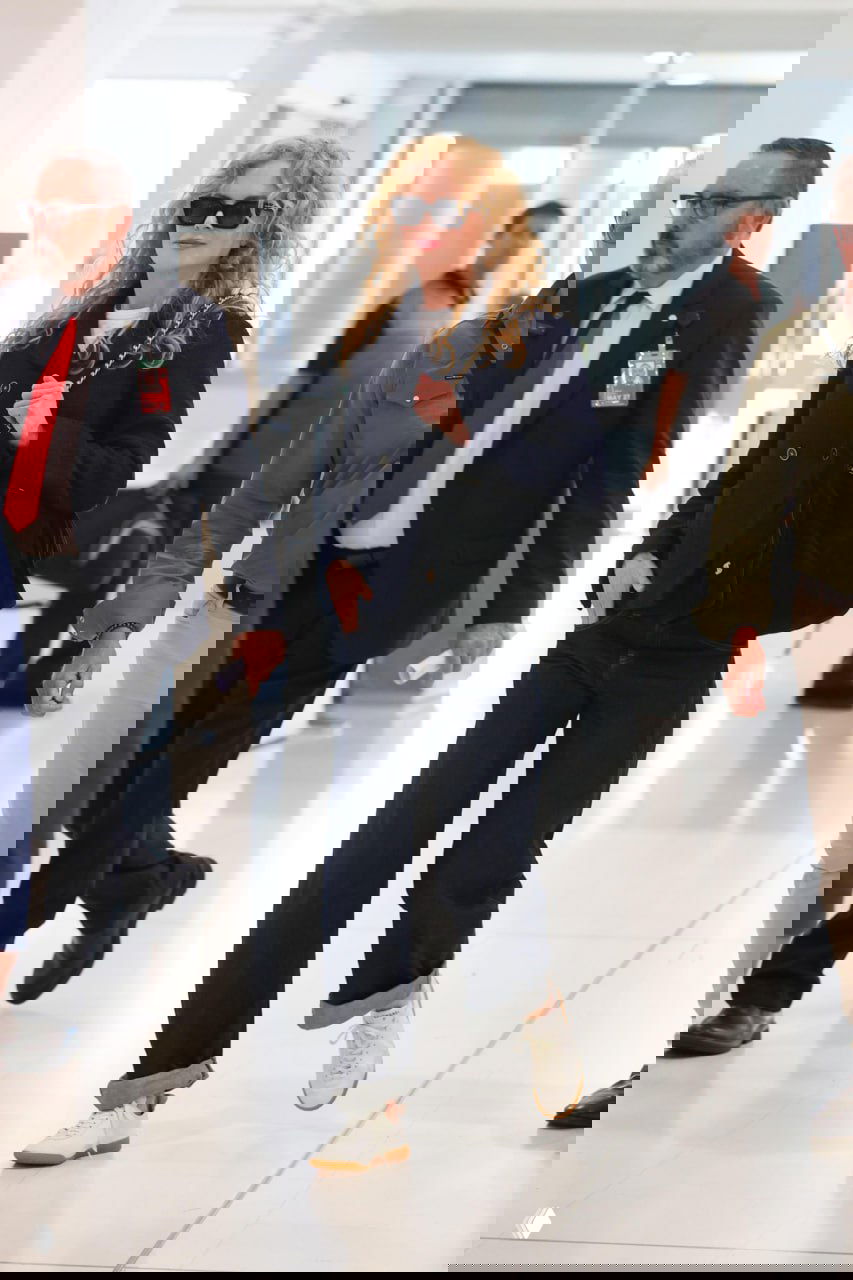 Женщина с кудрями в тёмном пальто и белых кроссовках идёт по терминалу аэровокзала в солнцезащитных очках. Фотография уличного папарацци.