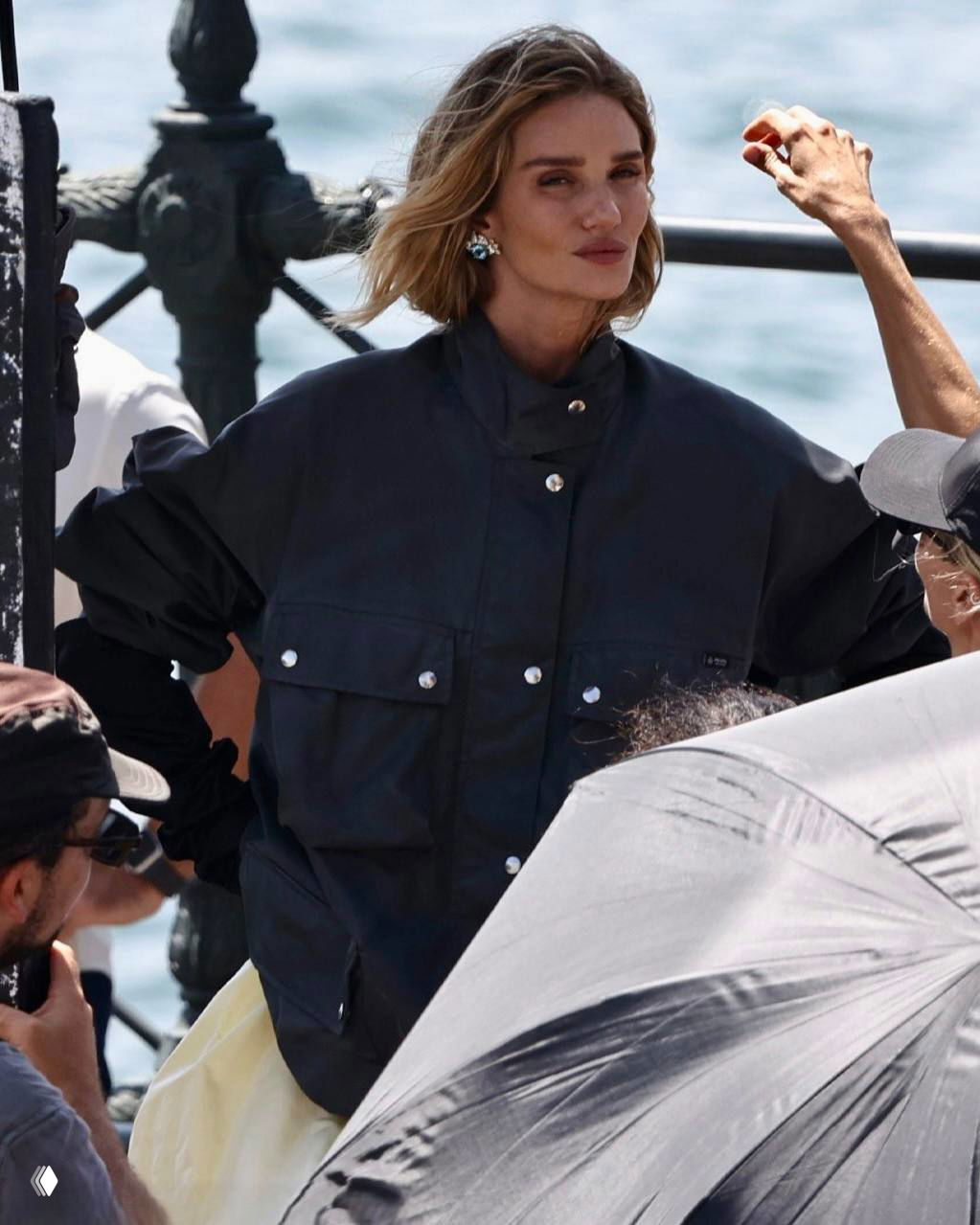 Роузи Хантингтон-Уайтли на набережной в тёмной куртке во время фэшн-съёмки в Сиднее; фотографы и осветители вокруг.