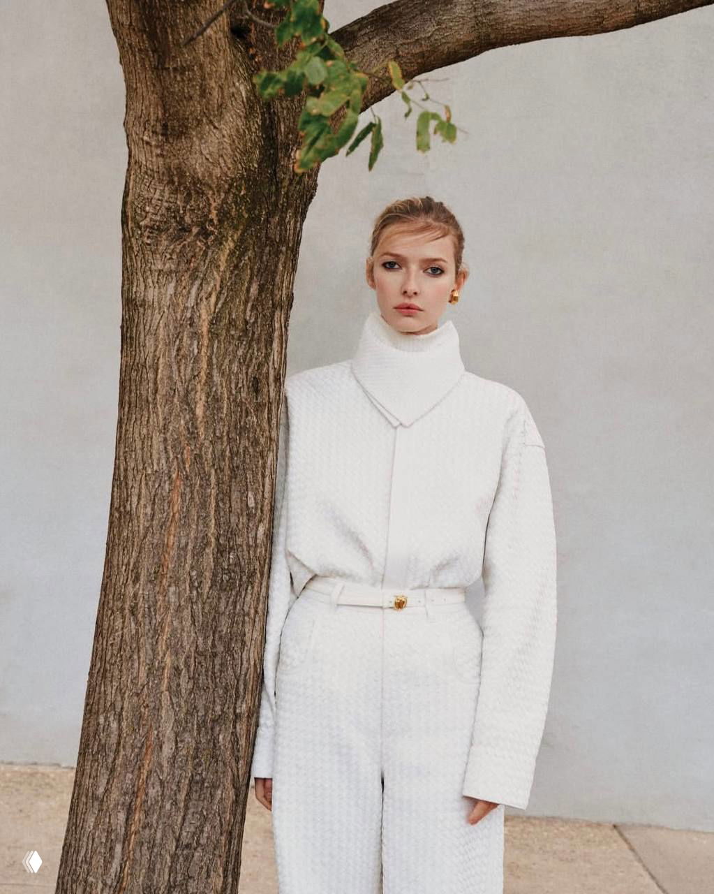 Женщина в белом стёганом комплекте стоит рядом со стволом дерева — контраст природного фона и минималистичного образа в редакционной съёмке.