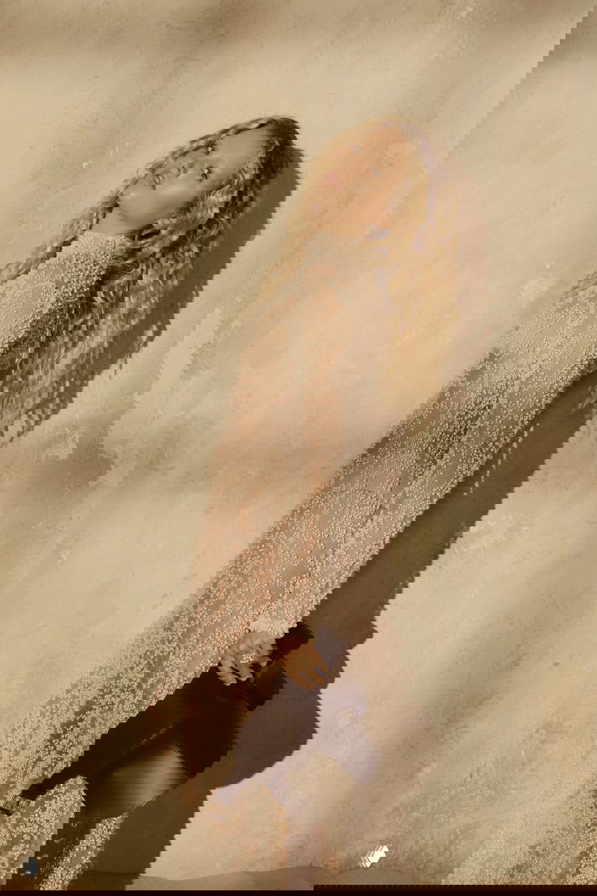 Модель стоит в полупрозрачном платье с декором-бусинами, поза в профиль на фоне текстурной стены и тёплое боковое освещение студийной съемки.