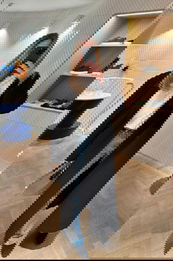 Зеркальное селфи: длинное чёрное пальто, белая футболка и светлые джинсы на фоне бутика — парижская классика для вытянутого силуэта.