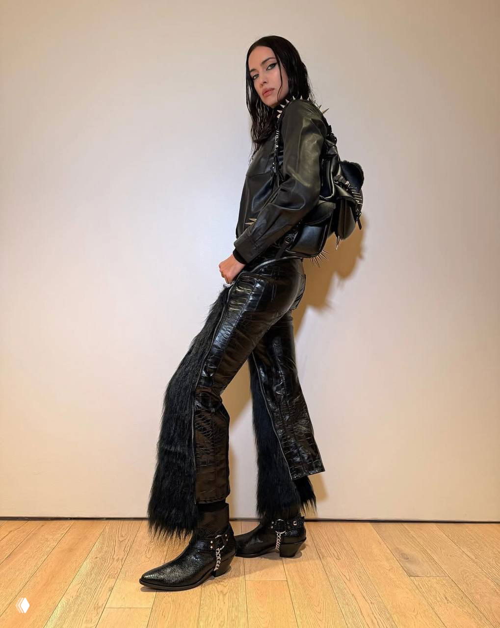 Полнотелый портрет модели в чёрном кожаном комплекте и ковбойских ботинках с цепочками на светлом фоне.