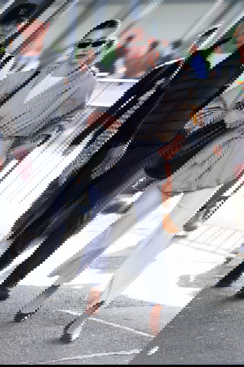 Молодая женщина в объёмной светлой куртке и белых брюках идёт по улице с наушниками и большой сумкой на плече, городской фон. Фотография уличного папарацци.