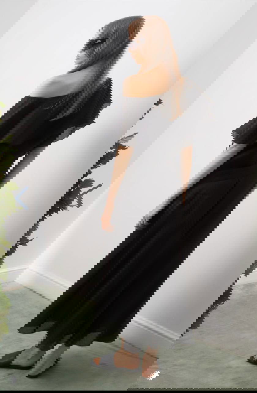 Модель в чёрном платье с открытыми плечами и широким воланом, вид со спины; лёгкая ткань, интерьерная съёмка в спокойной гамме и зелёными акцентами.