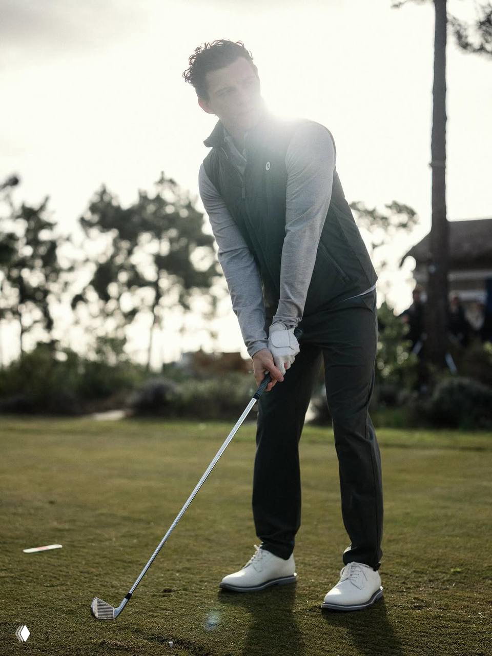 Мужчина в спортивной куртке и брюках готовится к удару клюшкой для гольфа на зелёном поле, съёмка подчёркивает активный образ жизни и одежду.