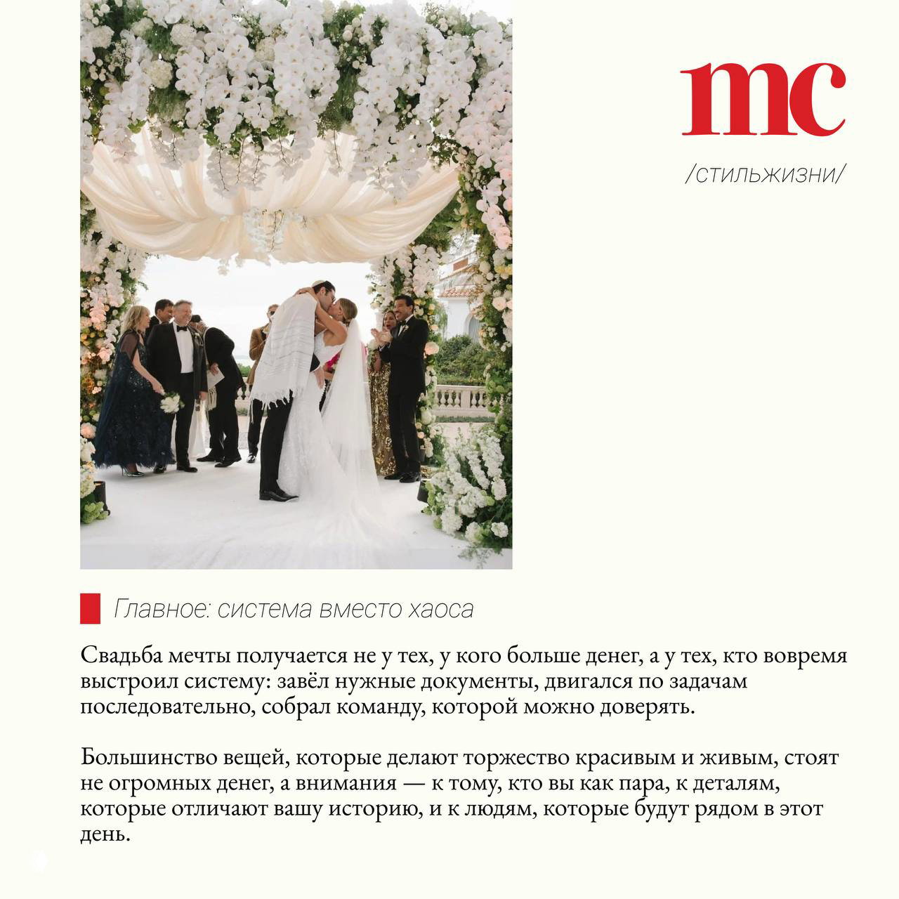 Свадебная церемония под аркой из белых цветов: жених и невеста целуются на фоне оформленной цветами сцены и приглашённых гостей.