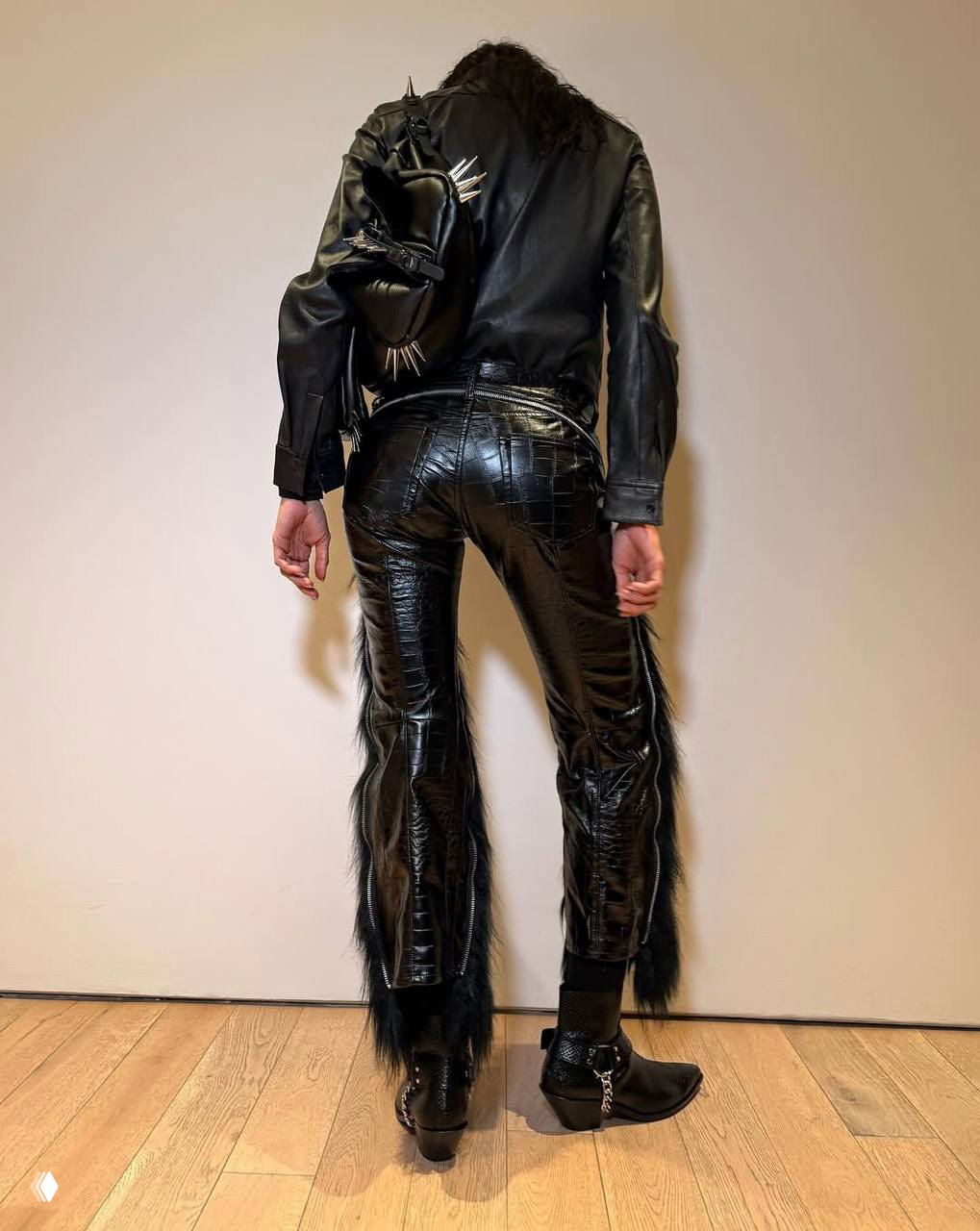 Кадр со спины: модель в узких кожаных брюках с фактурой и вставками меха, рюкзак со шипами виден сбоку.