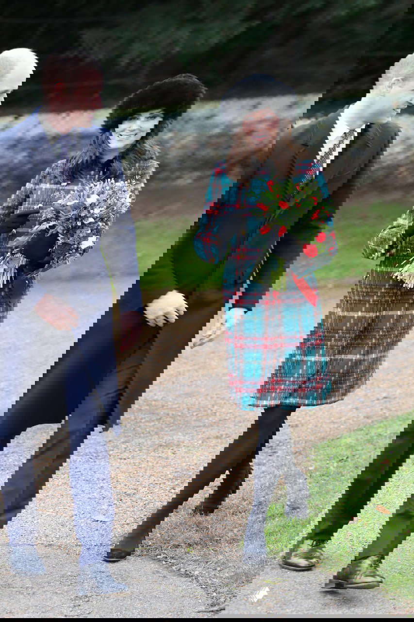 Женщина в тёплом клетчатом пальто и меховой шапке держит букет цветов, рядом мужчина в тёмном пальто — кадр с праздничной прогулки по парку.