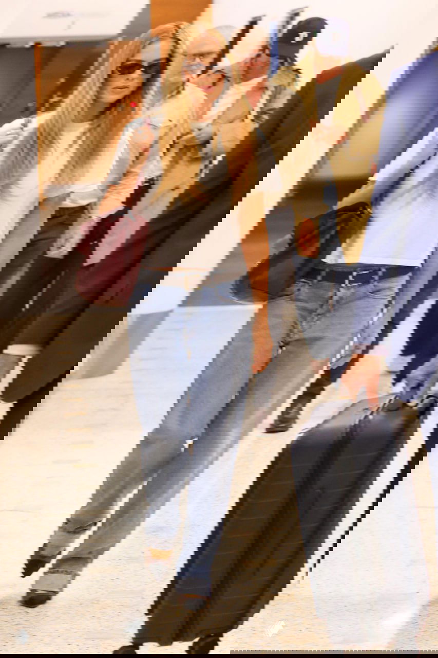 Женщина в свободной белой футболке и джинсах идёт по аэропортовому терминалу рядом с чемоданом, сумка на плече. Фотография уличного папарацци.