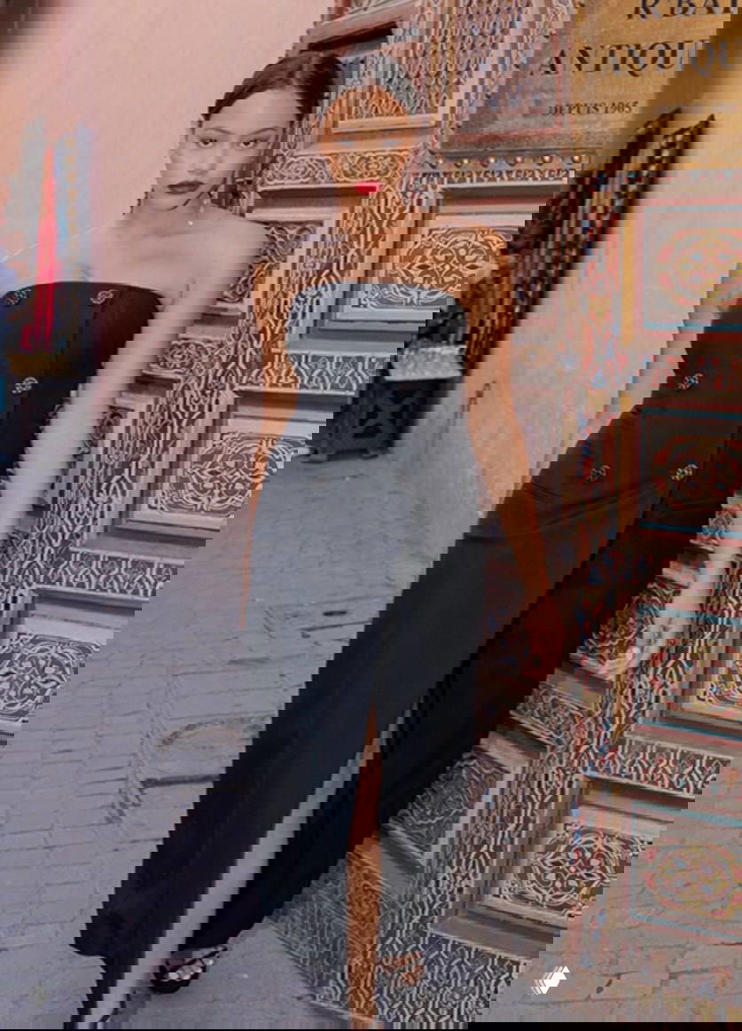 Уличная фотография: женщина в чёрном корсетном платье без бретелей с декоративными пуговицами и высоким разрезом, городской фон и узорчатая дверь рядом.