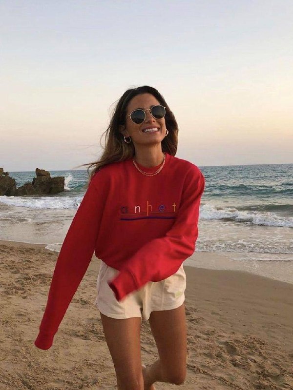 Женщина в красном свитере и солнцезащитных очках идет по пляжу на закате; море и скалы на заднем плане, расслабленная улыбка и естественный свет.