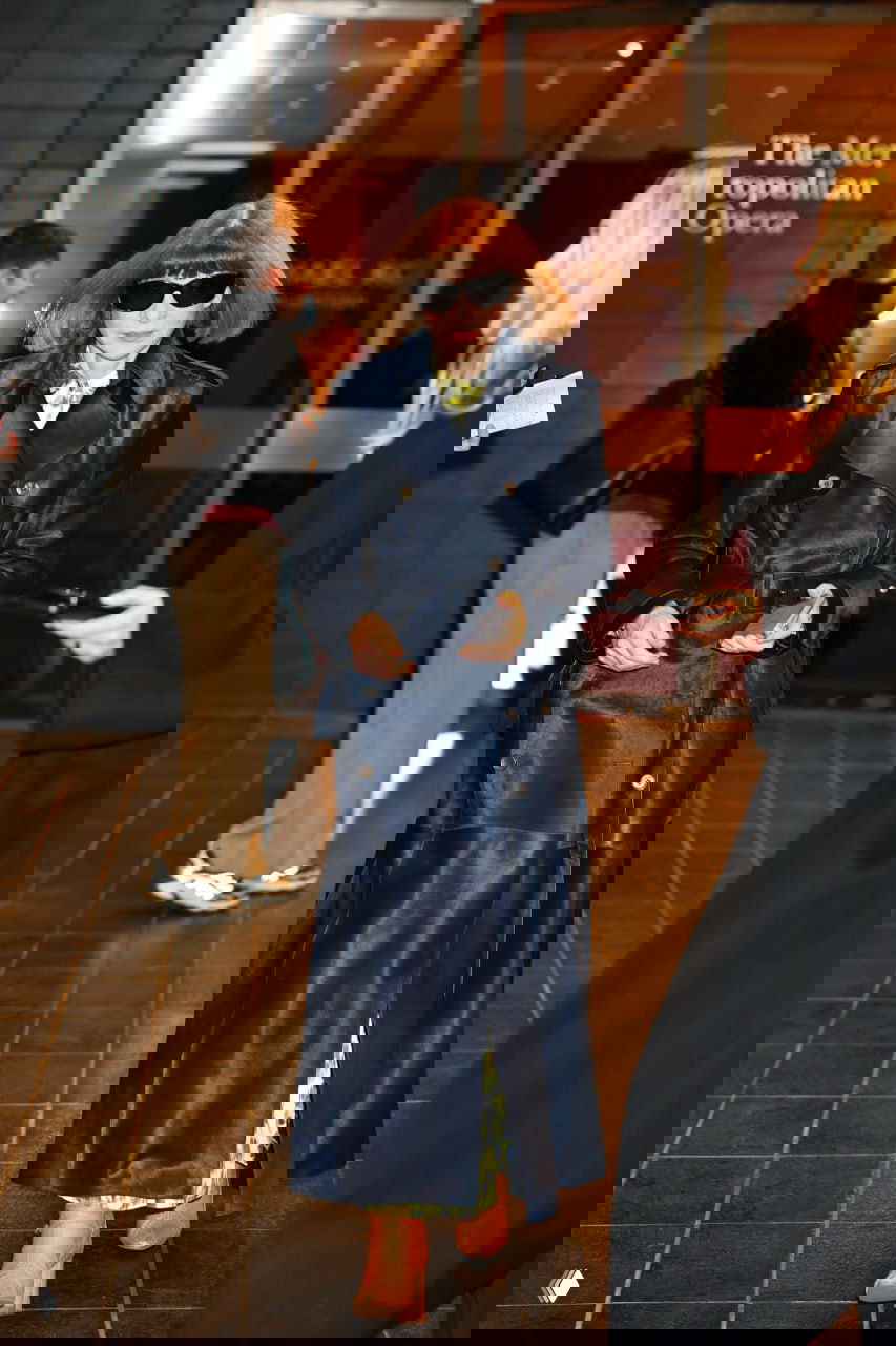 Пожилая женщина в длинном тёмном плаще и крупных солнцезащитных очках идёт у входа в здание театра, вечерняя городская сцена. Фотография уличного папарацци.