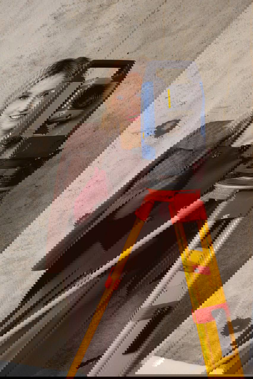 Женщина-геодезист с тахеометром на треноге рядом со строительной стеной; портрет на фоне рабочего оборудования.