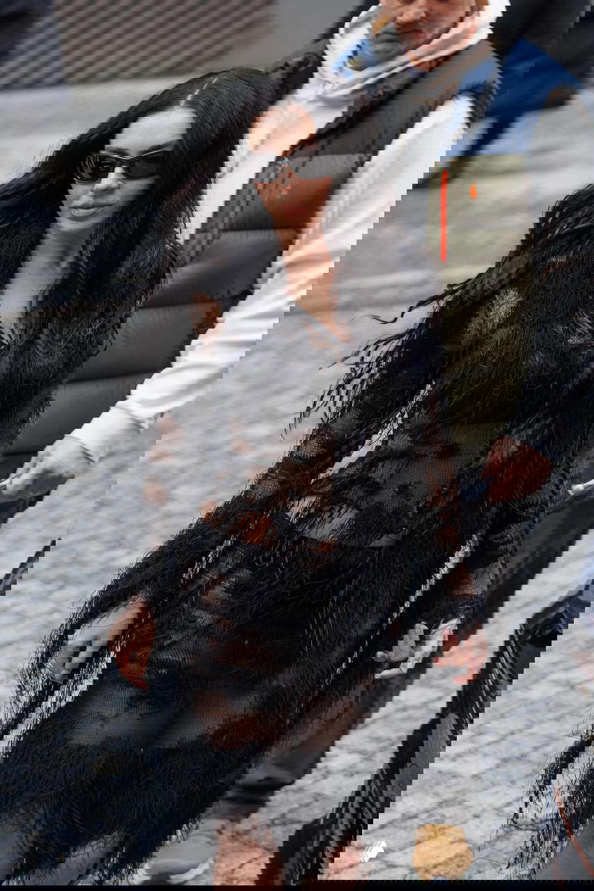 Длинное черное платье с полупрозрачными фрагментами и бахромой; длинные темные волосы, солнцезащитные очки и городской уличный антураж — гранж‑мастерская эстетика.