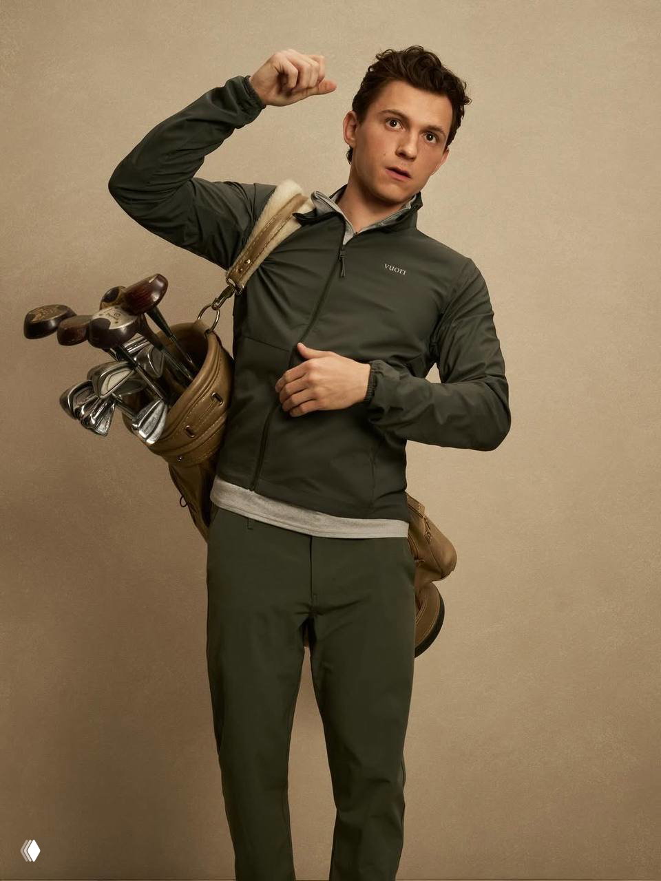 Фотография мужчины в тёмно-зелёной ветровке с гольф-сумкой за плечом в рекламной съёмке бренда спортивной одежды на нейтральном фоне.