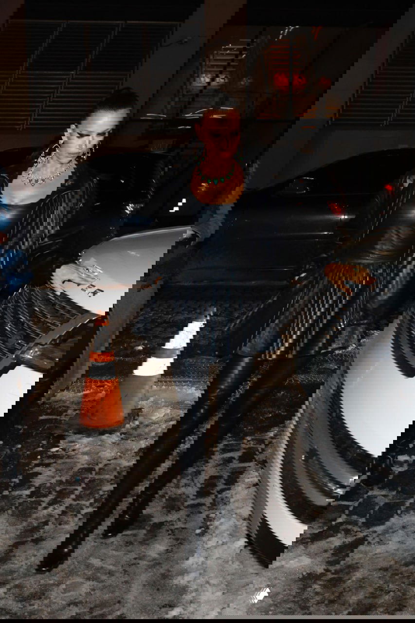 Женщина в чёрном кожаном мини-платье и высоких сапогах идёт по мокрой брусчатке рядом с припаркованным автомобилем, ночная сцена. Фотография уличного папарацци.