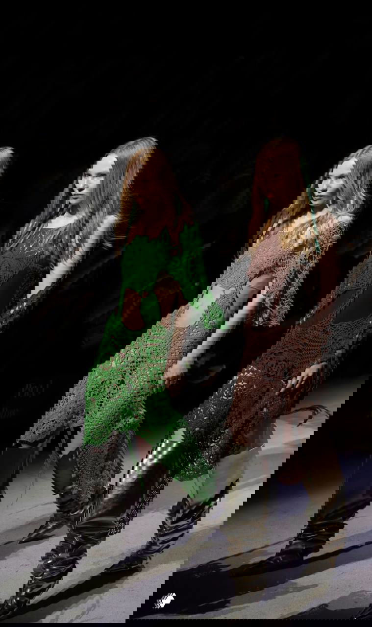 Показ на подиуме: модели в вязаных платьях зелёного и коричневого оттенков с вырезами и высокими блестящими сапогами, драматическое освещение сцены.