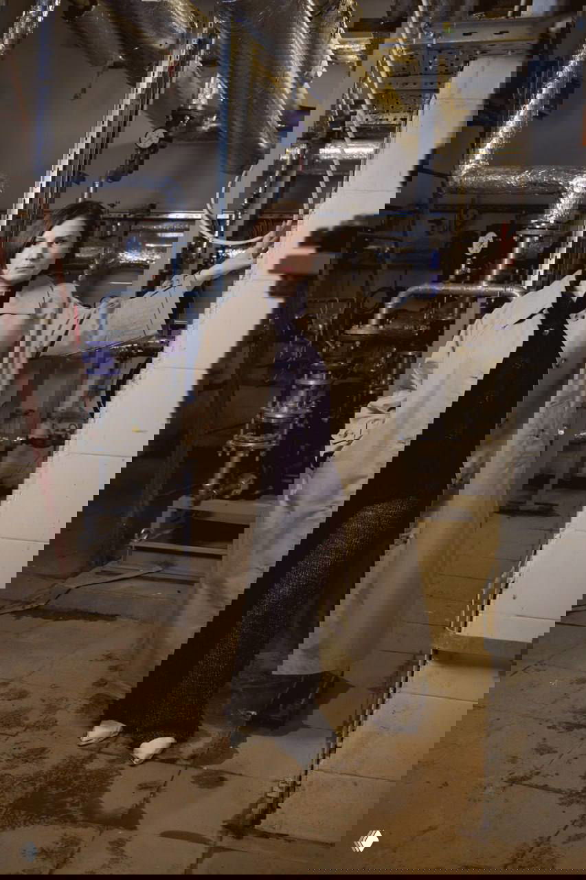 Женщина в длинном плаще в техническом помещении с трубами и инженерным оборудованием за спиной, рабочая среда инженерных сетей.