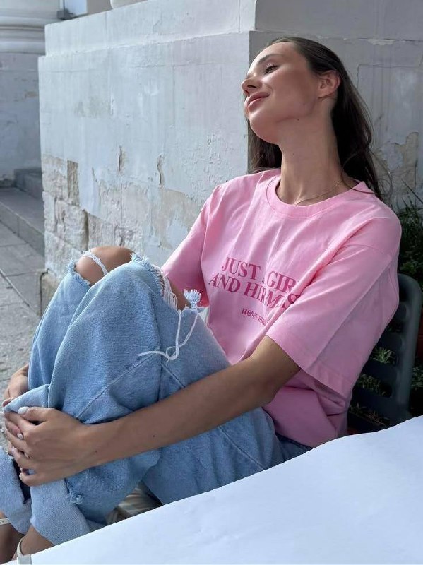 Портрет молодой женщины в розовой футболке и светлых рваных джинсах, сидит у стены и наслаждается моментом; мягкое дневное освещение и спокойная поза.