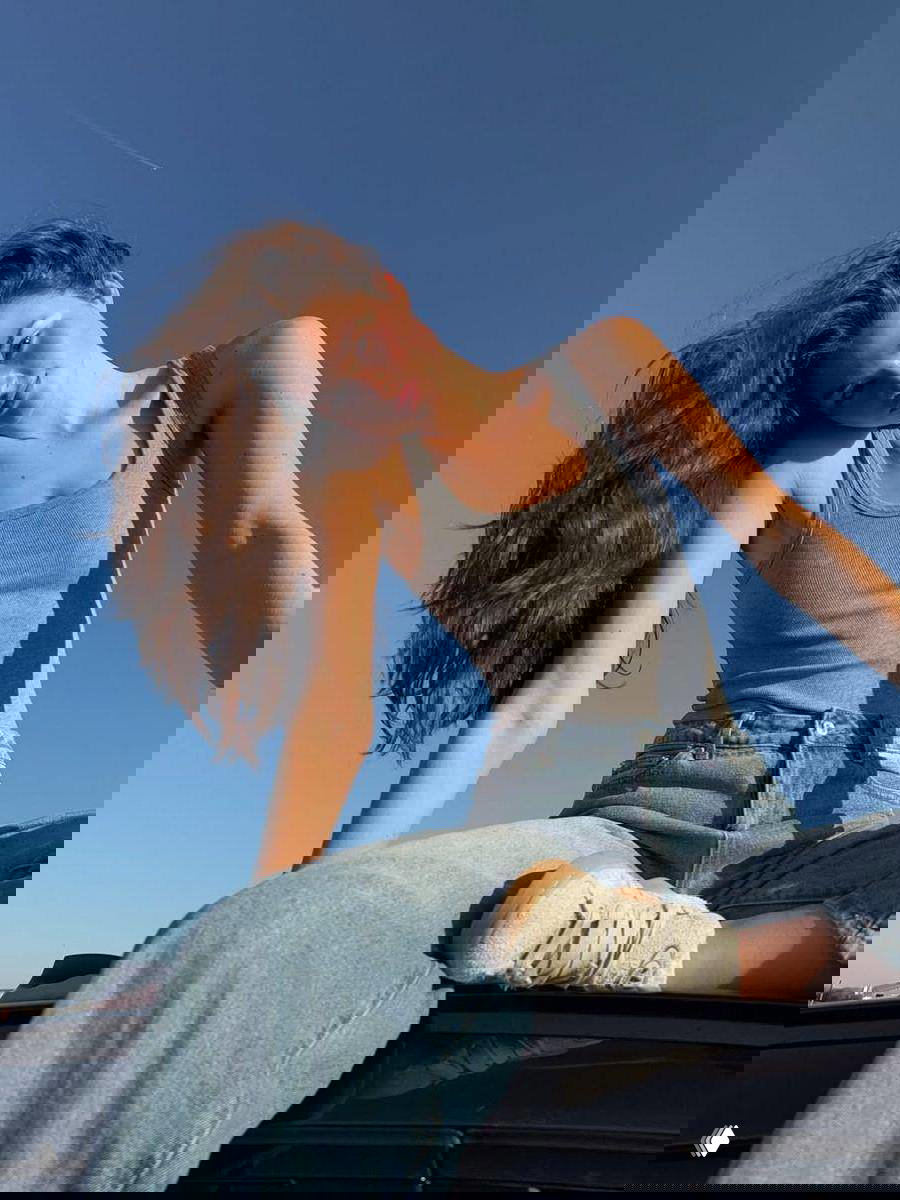 Девушка в сером топе и джинсах позирует на автомобиле под чистым синим небом; солнце подсвечивает волосы, летняя непринужденная сцена.