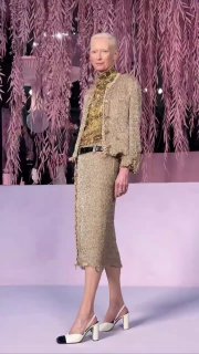 Высокая бледная модель идет по подиуму в бежевом твидовом костюме и белых туфлях; минималистичный макияж и строгая посадка образа на показе Chanel.