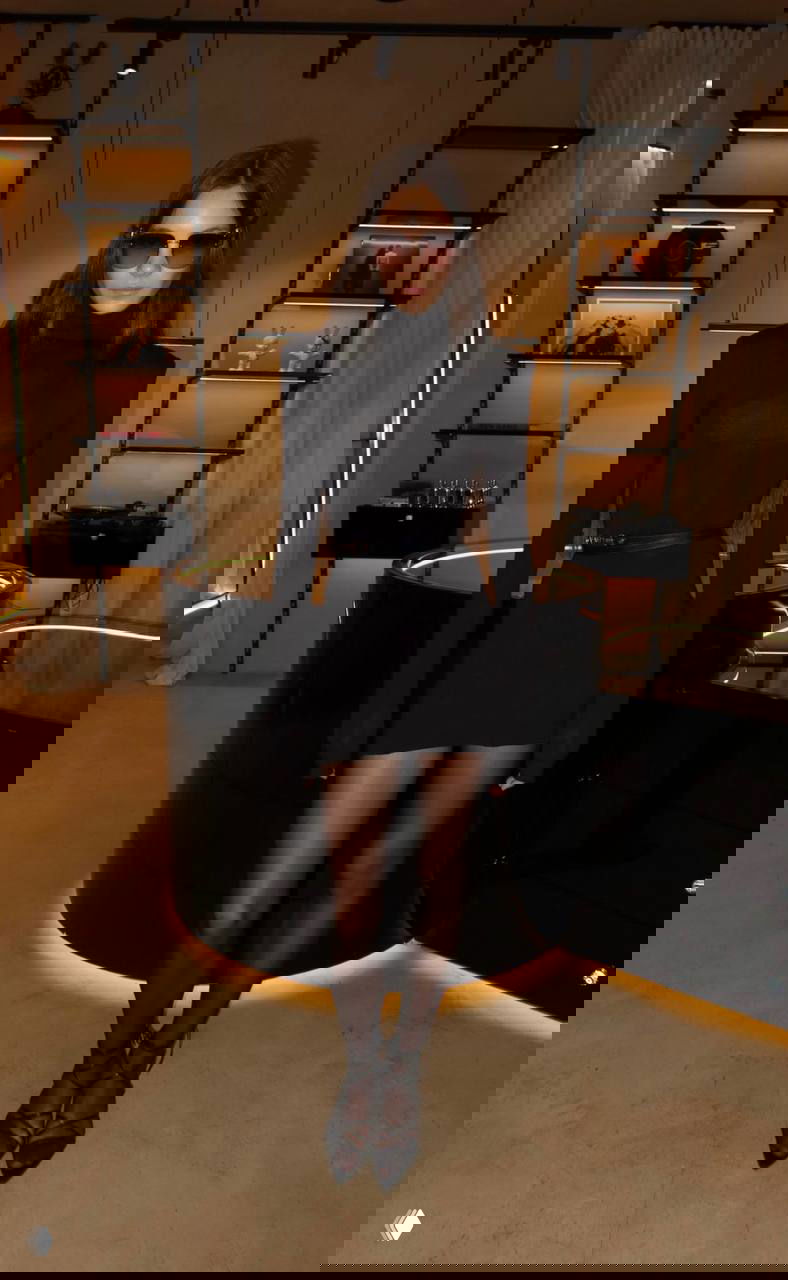 Интерьерная ночная фотография: модель в чёрном мини‑платье с высоким воротником‑стойкой и солнцезащитными очками, атмосфера клуба или лобби отеля.