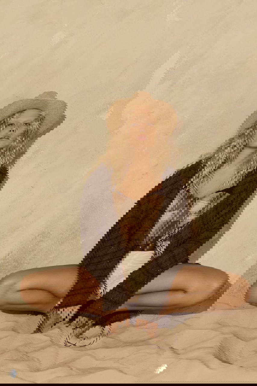 Модель сидит в шляпе и свободной куртке на песке, кадр с центральной композицией и естественной позой, передающей расслабленное летнее настроение съемки.