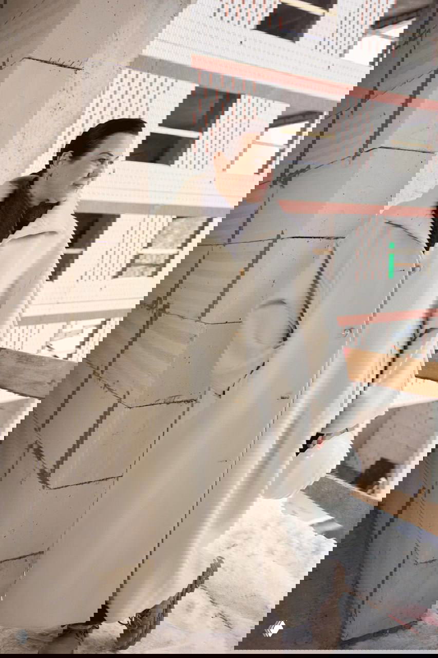 Женщина в светлом пальто и с каской в руках стоит на балконе строящегося дома; видны кирпичные стены и проёмы.