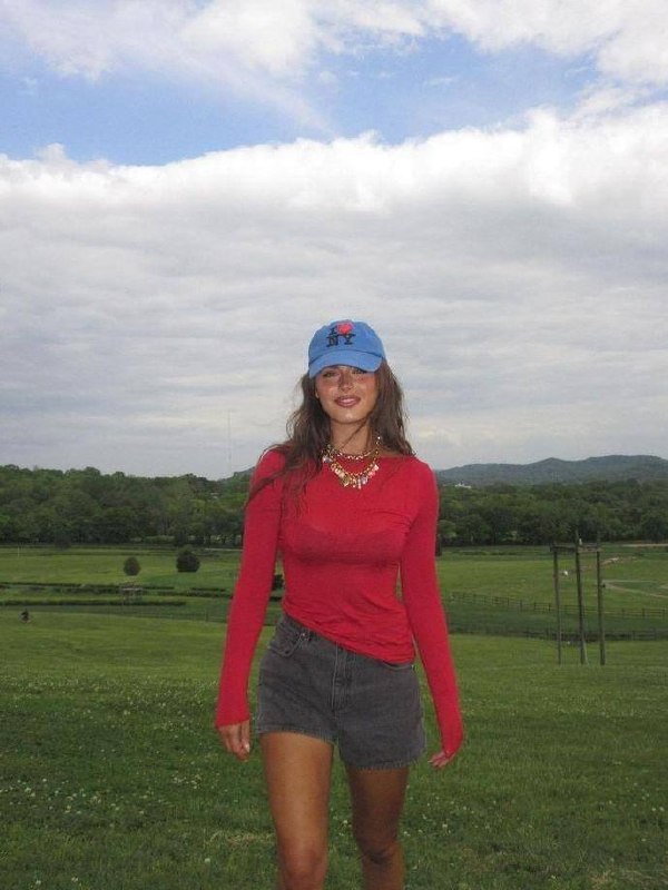 Молодая женщина в красном свитере и голубой кепке идет по зеленому полю на фоне холмов и облачного неба; образ в стиле casual и природа вокруг.