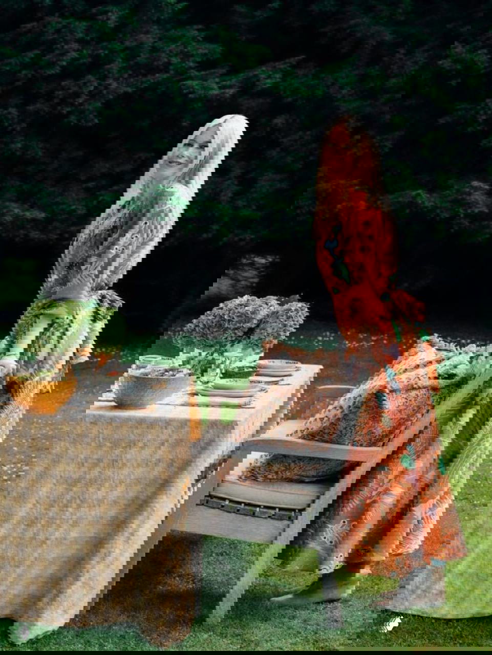 Женщина в длинном платье рядом с сервированным столом на газоне; весенняя сервировка и природный зелёный фон подчёркивают загородную атмосферу праздника.