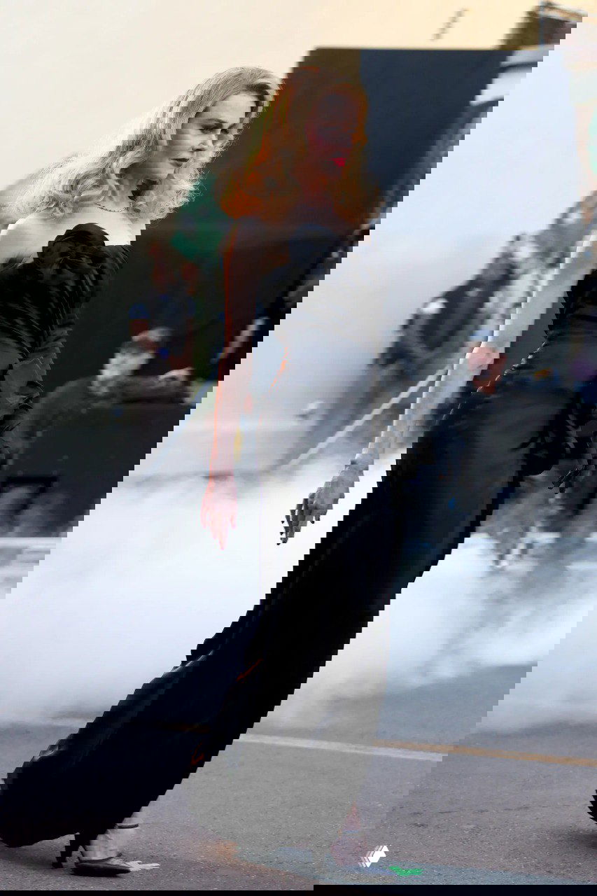 Профиль актрисы в длинном чёрном платье Chanel: стройный силуэт, волнистые светлые волосы и тонкий вечерний макияж на фоне съёмочной площадки.