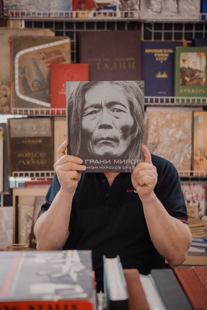 Продавец в книжном павильоне держит перед лицом крупную книжную обложку с портретом, вокруг — стопки старых книг и плакаты.