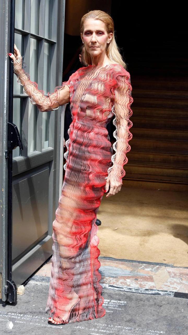 Селин в облегающем красно‑персиковом платье с волнистой фактурой ткани у дверного проёма — современный прогулочный образ и элегантность силуэта.