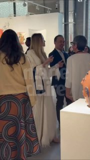 Женщина в светлом пальто среди посетителей в галерее на открытии Арт‑Базель в Дохе; видны картины и люди, обсуждающие экспонаты.