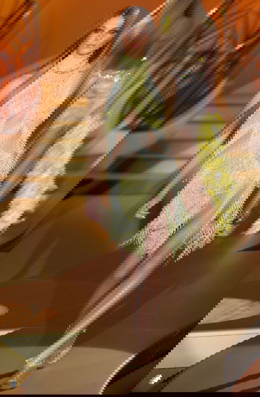 Хрупкая модель в зеленовато-жёлтом ажурном платье с бахромой и прозрачными элементами Versace 2004 идёт по подиуму с плавными разрезами и струящейся бахромой.